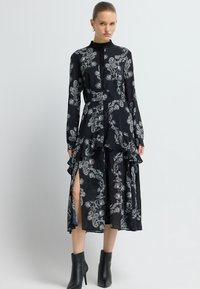 Zwarte lange mouwen jurk met een wit paisleypatroon, ruches aan de onderkant, hoge halslijn en zijsplitten; gecombineerd met zwarte enkellaarsjes.