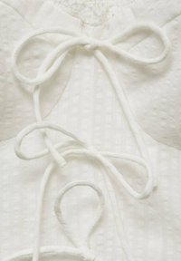 Tissu blanc avec des rayures texturées, doté d'une fermeture à nœud papillon proéminente et d'accents arrondis. Le design est simple et minimaliste.