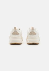 White athletic shoes with a textured mesh heel and beige accents. Features a padded collar and a rubber sole with strategic traction.