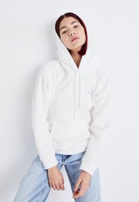 Vit hoodie med känguruficka, justerbar huva och subtil logotypdetalj på bröstet, tillsammans med ljusblå jeans. Slät yta.