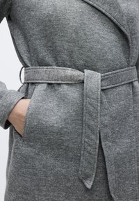 Γκρι παλτό με υφασμάτινη υφή, ζώνη στη μέση και πλαϊνές τσέπες. Ο γιακάς είναι σε στιλ κασκόλ, προσδίδοντας συνολικά μια ομαλή σχεδίαση.