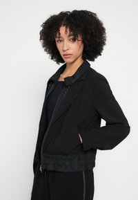 Geacă bomber neagră ușoară, cu fermoar, manșete elastice și talie strânsă. Are o textură fină și un design minimalist.