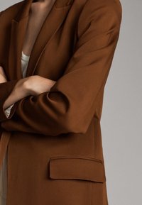 Brązowa, dopasowana marynarka o usztywnionej konstrukcji, z klapą z wycięciem, pojedynczą kieszenią oraz gładką, miękką fakturą tkaniny.