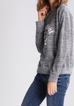 Kvinde iført en grå langærmet sweater med hvid tekst og blå jeans, stående med den ene hånd nær ansigtet og den anden ved siden af.