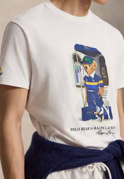 Λευκό t-shirt με εκτυπωμένη αρκούδα σε μπλε και κίτρινο polo outfit που κρατάει μια σημαία και μια τσάντα, πουλόβερ δεμένο στη μέση, κουτσουρεμένη άποψη του πάνω μέρους του σώματος.