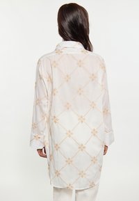 Witte blouse met lange mouwen en een open kraag. Bevat een geborduurd ruitpatroon in lichtbruin, met een lichte,Doorschijnende stofstructuur.