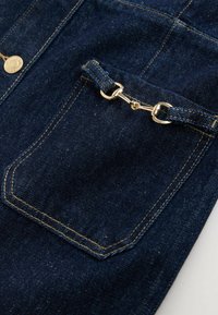 Poche en denim bleu foncé avec détail en chaîne dorée et coutures couleur tabac sur tissu en denim, avec un bouton en laiton visible près du coin supérieur gauche.