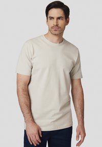 Beżowa bawełniana koszulka z okrągłym dekoltem i krótkimi rękawami, zaprezentowana na modelu, zestawiona z ciemnymi spodniami, prosty design, bez wzorów.