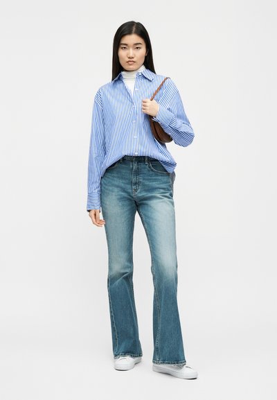 Νεαρή γυναίκα που φοράει μπλε και άσπρη ριγέ μπλούζα, λευκό ζιβάγκο, ξεθωριαμένα μπλε τζιν, λευκά αθλητικά και κρατάει καφέ τσάντα ώμου.