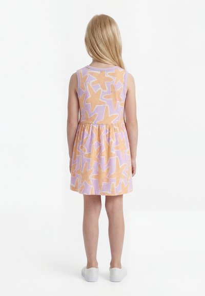 Κορίτσι με μακριά ξανθά μαλλιά, που φοράει ροζ αμάνικο φόρεμα με μεγάλα πορτοκαλί αστέρια, στέκεται με την πλάτη προς το μέρος μας, λευκά παπούτσια.