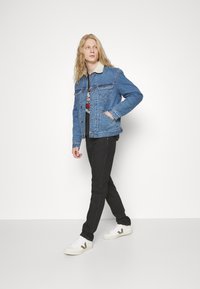 Giacca di jeans con lavaggio blu chiaro, colletto a contrasto, tasche frontali, indossata su una t-shirt grafica nera, abbinata a pantaloni neri e sneakers bianche.