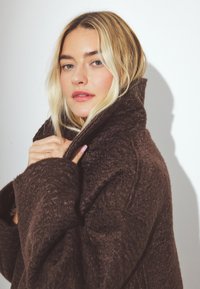 Brązowy, strukturalny płaszcz z wysokim kołnierzem, grubego materiału i oversize'owym kroju. Modelka trzyma kołnierz, podkreślając jego ciepło i design.