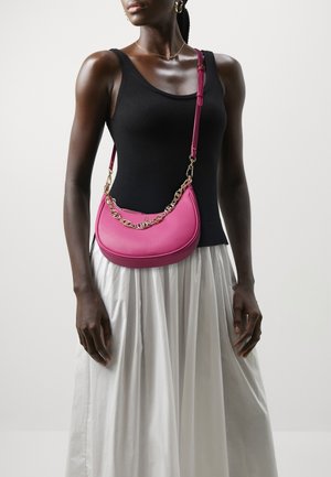 Roze leren crossbodytas met een gebogen vorm, gouden kettingaccent en verstelbare band. Draagt een zwarte top en een witte rok.