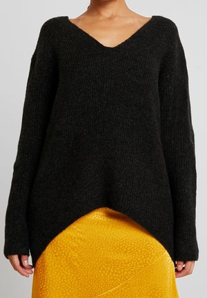 Woman wearing a textured black V-neck sweater with long sleeves and a yellow patterned skirt with a wrap detail.
