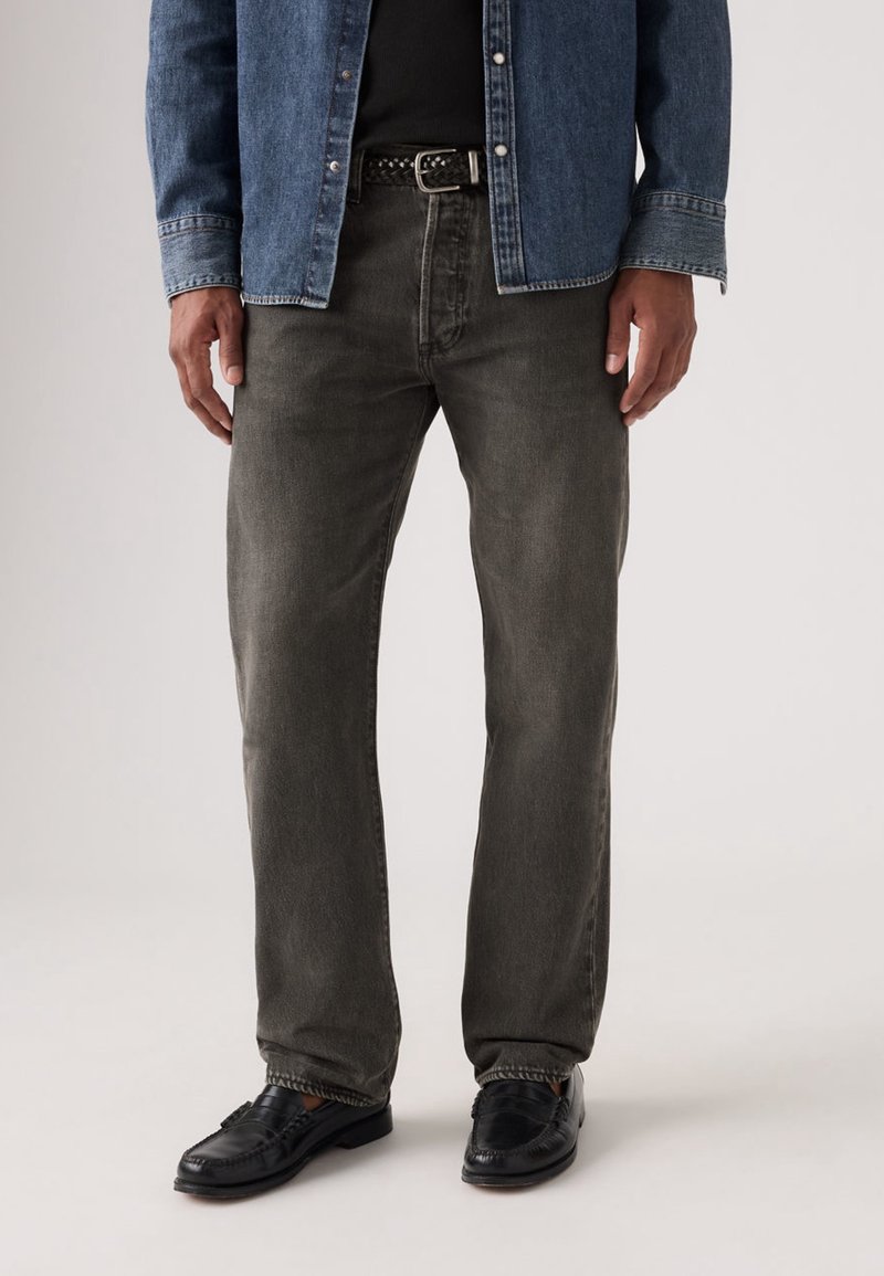Homme portant un jean droit gris foncé, une ceinture noire, des chaussures noires et une chemise en denim bleue partiellement boutonnée par-dessus un top noir.