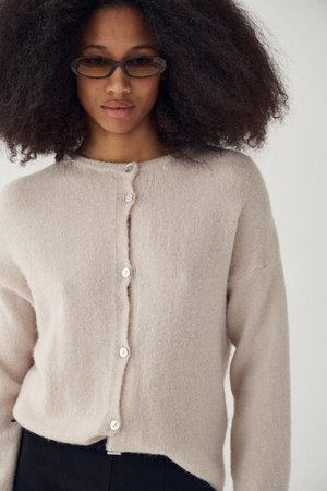 Mlada ženska z velikimi kodrastimi lasmi, ki nosi ovalna sončna očala jantarne barve in bež cardigan z gumbi ter črne hlače.