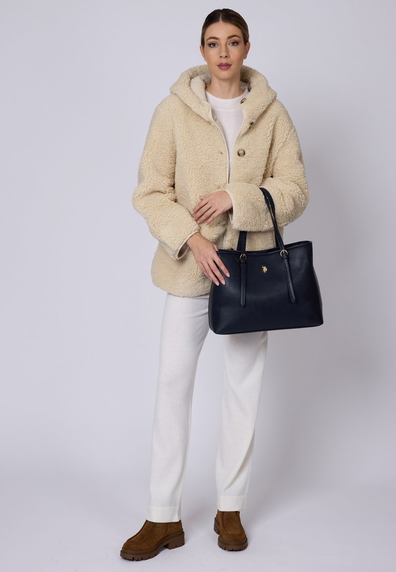 Beżowa puszysta kurtka z kapturem, biała sweter, kremowe spodnie, brązowe botki za kostkę oraz granatowa skórzana torba na ramię z złotymi detalami.
