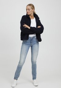 Marineblå fleece zip-up jakke med høy krage, kombinert med lyseblå skinny jeans og hvite joggesko, vist mot en nøytral bakgrunn.