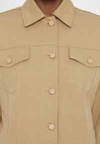 Beige denim-takki, jossa on kauluksellinen kaula-aukko, viisi nappia, kaksi rintataskua ja näkyvät ompeluyksityiskohdat. Kankaan rakenne on teksturoitu ja istuvuus on muotoiltu.