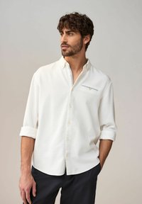 Chemise blanche à boutons, avec un motif texturé, manches longues retroussées, et un petit détail de poche décoratif avec un accent bleu sur la poitrine.