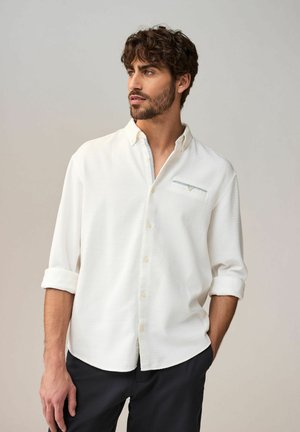 Chemise blanche à boutons, avec un motif texturé, manches longues retroussées, et un petit détail de poche décoratif avec un accent bleu sur la poitrine.