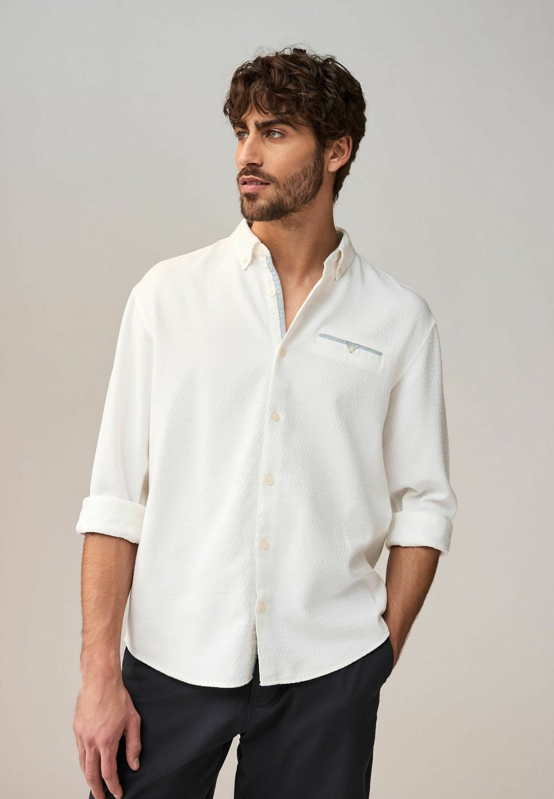 Chemise blanche à boutons, avec un motif texturé, manches longues retroussées, et un petit détail de poche décoratif avec un accent bleu sur la poitrine.