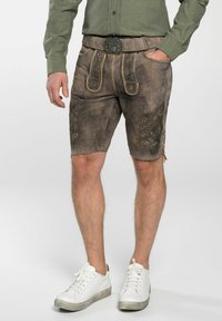 Braune Lederhosen mit gelben Nähten, dekorativer Stickerei und einem Gürtel mit einer Metallschnalle. Getragen mit weißen Turnschuhen.