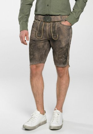 Braune Lederhosen mit gelben Nähten, dekorativer Stickerei und einem Gürtel mit einer Metallschnalle. Getragen mit weißen Turnschuhen.