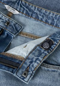 Le tissu en denim bleu présente une fermeture éclair, des coutures apparentes, une fermeture à bouton et une doublure intérieure douce avec une poche.