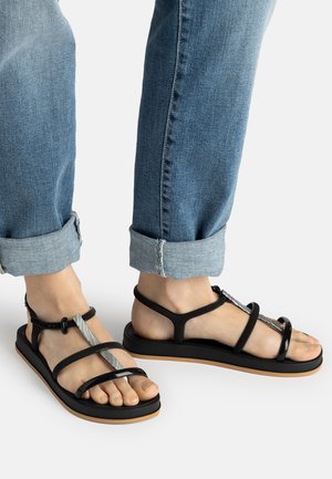 Person, die hochgekrempelte blaue Jeans und schwarze flache Sandalen mit mehreren dünnen Riemen und einer hellbraunen Sohle vor weißem Hintergrund trägt.