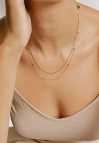 Collane in oro con strati sottili e piccoli dettagli di perline, appoggiate su uno sfondo di tessuto liscio e neutro.