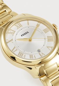 Montre Fossil dorée avec un boîtier rond, un cadran argenté, des chiffres romains, un affichage de la date et un bracelet à maillons. Fini métallique lisse sur l'ensemble.