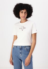 Cremefarvet T-shirt med korte ærmer, der har "New York Tommy Hilfiger" logo i bordeaux og et lille logo detajle på ærmet. Båret med blå jeans.