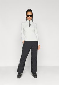 Pulóver blanco de manga larga con medio cierre y pantalones negros con franjas laterales. El modelo lleva gafas de sol negras y botas de esquí. Texturas suaves.