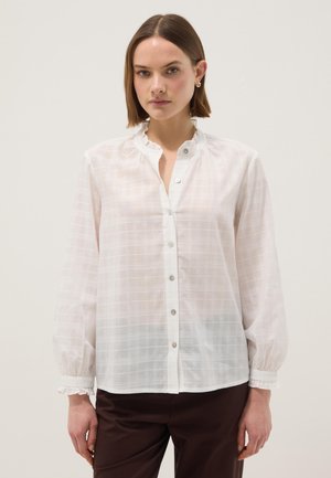 Femme portant un chemisier à carreaux blanc semi-transparent avec col et poignets volantés, pantalon marron et boucles d'oreilles anneaux dorés, se tenant devant un fond uni.