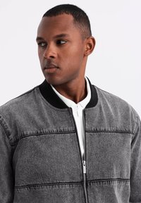 Giacca bomber grigia con colletto nero e zip bianca. Presenta cuciture orizzontali e una texture simile al denim. Design semplice e moderno.