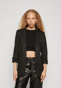 Jeune personne aux longs cheveux bouclés porte un blazer noir scintillant, un top noir court et un pantalon en cuir noir sur fond clair.