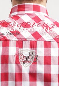 Camicia a scacchi rossa e bianca con colletto alla coreana, caratterizzata da dettagli ricamati, una toppa a scudo e cuciture decorative sul retro.