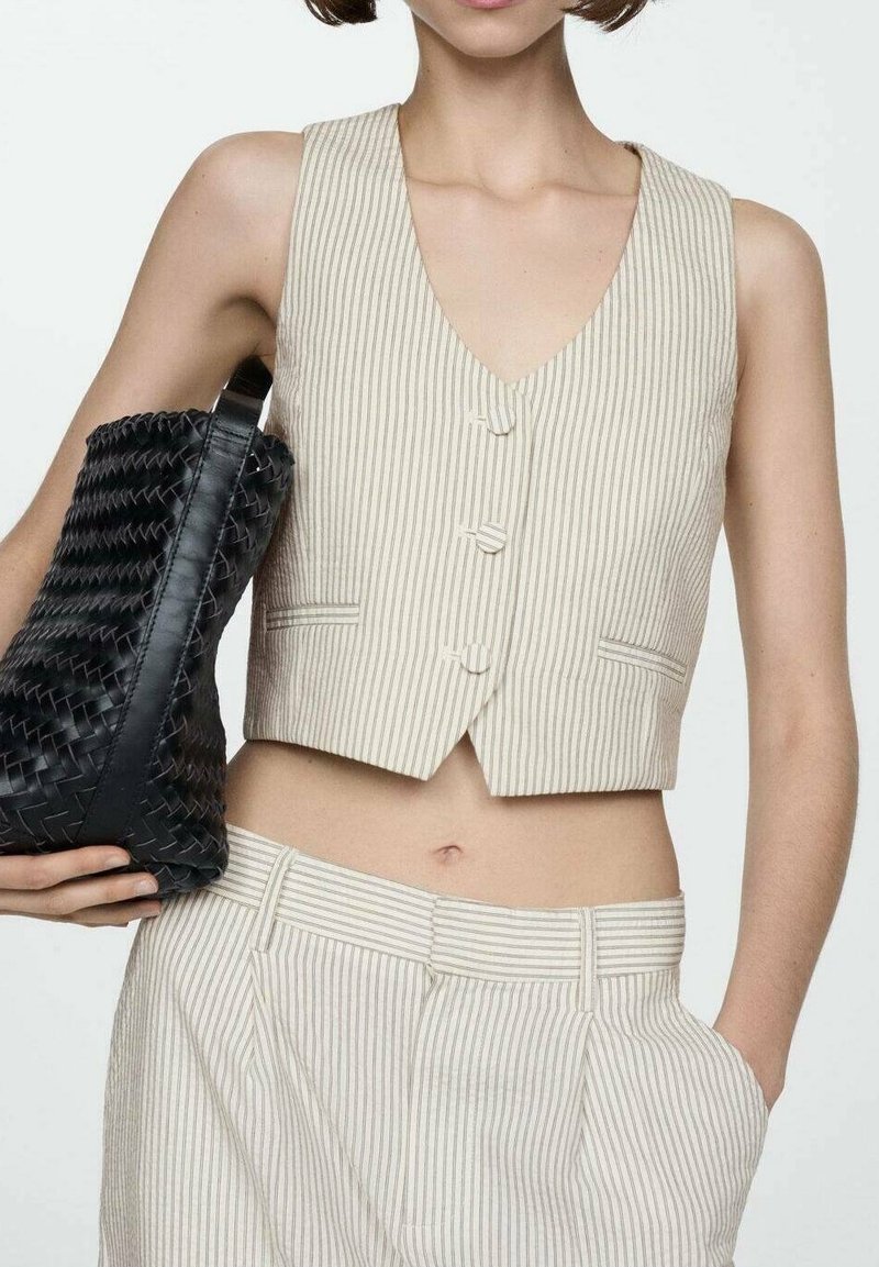 Femme portant un gilet sans manches beige à fines rayures et un pantalon assorti, tenant un grand sac à main noir tissé sous le bras.