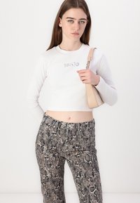 Valkoinen lyhyt hihainen toppi, jossa on "HUGO"-logo; kuvioidut käärmeennahkakuosiset housut eri harmaan ja mustan sävyissä; beige käsilaukku olkahihnalla.