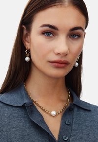 Een gouden sieradenset met witte parels op de oorbellen en een ketting. Het model draagt een grijze trui met kraag.