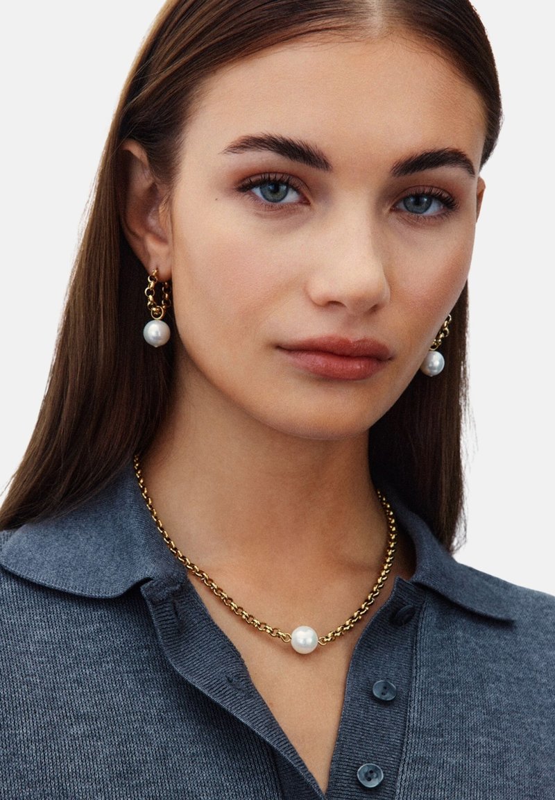 Een gouden sieradenset met witte parels op de oorbellen en een ketting. Het model draagt een grijze trui met kraag.