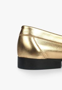 Chaussure slip-on en métal doré avec un dessus en cuir lisse, forme arrondie et une semelle en caoutchouc noire. Présente des accents de couture subtils.