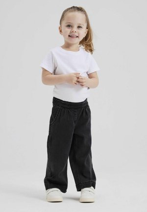 Pantaloni in denim neri con cintura elastica, abbinati a una maglietta bianca a maniche corte e sneakers bianche. Bambino in piedi, sorridente.
