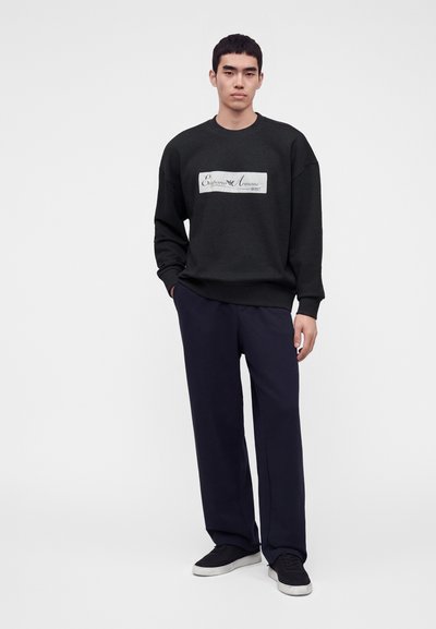 Νέος άνδρας που φοράει μαύρο φούτερ Emporio Armani, σκούρο φαρδύ παντελόνι και μαύρα αθλητικά παπούτσια με λευκές σόλες, στέκεται μπροστά σε απλό φόντο.