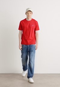 T-shirt rouge à manches courtes avec une poche poitrine ; associé à un jean bleu délavé et des baskets blanches ; modèle portant une casquette claire.