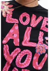 Černé bavlněné tričko s velkým růžovým nápisem s různými vzory a texturami, ozdobené květinovým motivem na levé straně.