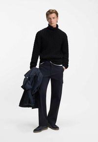 Schwarzer, grob gestrickter Rollkragenpullover, kombiniert mit dunkelblauen, weit geschnittenen Hosen. Das Model hält eine dunkle Jacke und trägt dunkle Schuhe mit strukturierten Sohlen.