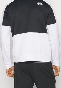 Sudadera deportiva de manga larga en blanco y negro con textura acanalada, que presenta un logo en la parte superior de la espalda y un diseño de paneles en contraste.