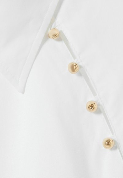 Primo piano di una camicia bianca con colletto e tre bottoni dorati lungo la patta, che evidenzia un design pulito ed elegante.